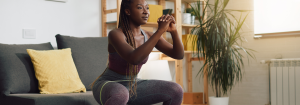 A young black woman in sportswear doing a squat with good form over a mat at home in front of her sofa to represent improving fitness levels. A water bottle is in front of her and a green plant is in the background.