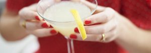 A lady with red nail varnish holding a cocktail with a slice of lemon in it.