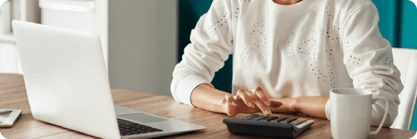 Setting a budget can help you to stop drinking alcohol. Image shows woman sat at a table in front of a computer typing on a calculator. 