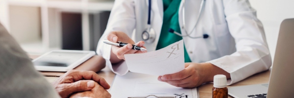 A doctor in a white coat at their desk looking at a piece of paper facing a patient.