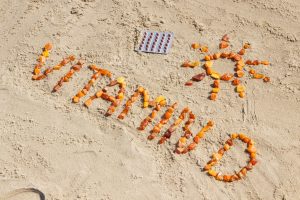 Vitamin D written in sand using orange stones next to some supplements with a sun shape next to it