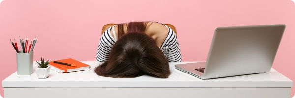 A woman sits with her head on the desk, a laptop is by her side.
