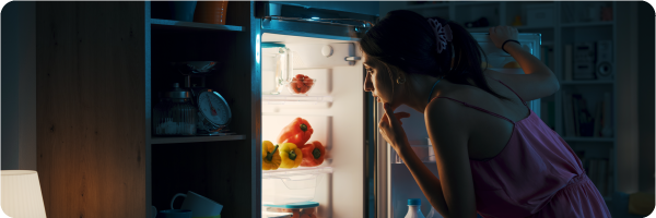 Avoid eating later for better sleep. Image shows a woman in nightwear looking in her fridge at night.