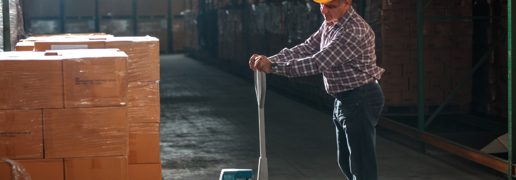 A slightly overweight middle aged man wearing a hardhat working a night shift in a dark warehouse moving boxes onto a palette.