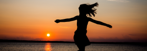 A silhouette of a young girl in a short sleeved t-shirt and skirt with her arms out and hair flying in the wind against an orange sky at sunset to illustrate the heath benefits of natural light. The sea can be seen in the background.