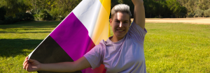 A non-binary person smiling holding the Non-binary pride flag standing on some gras to illsutrate how to observe International Non-Binary People's Day