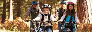 A Caucasian family, a man and a woman in the background and a young smiling boy and girl in front wearing helmets and cycling through a wooded area to represent the benefits of cycling