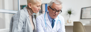 A middle aged Caucasian female patient sat with an older Caucasian male doctor as he supportively explains something to her.