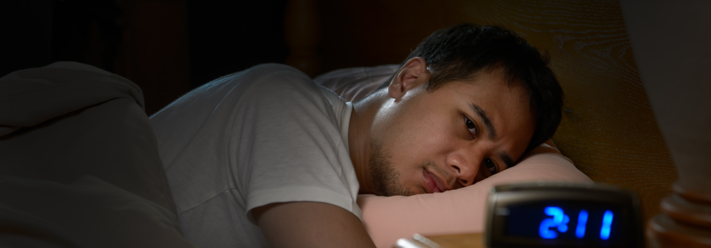 A man wearing a white t-shirt is lying in bed awake. His digital alarm clock reads 2:11.