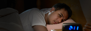 A man wearing a white t-shirt is lying in bed awake. His digital alarm clock reads 2:11.