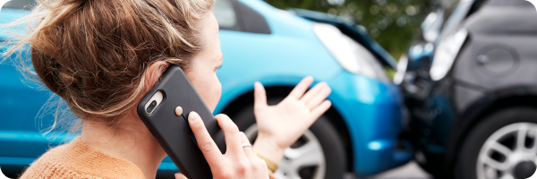 In the foreground a blonde-haired women is on a phone call. In the background the front of a blue car has collided with the back of a black car. There is damage to the bonnet.