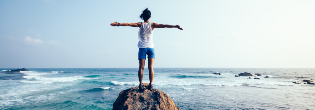 A young woman standing on a rock with her arms open looking out at rolling blue waves and sky.