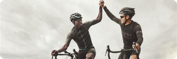 A young black man and a young Caucasian man wearing cycling helmets high fiving each other and smiling. Both are on bikes with a cloudy sky in the background. 