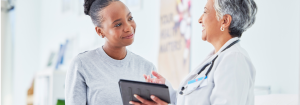 A smiling black woman talking to a smiling older Asian doctor wearing a white lab coat and stethoscope holding a tablet.
