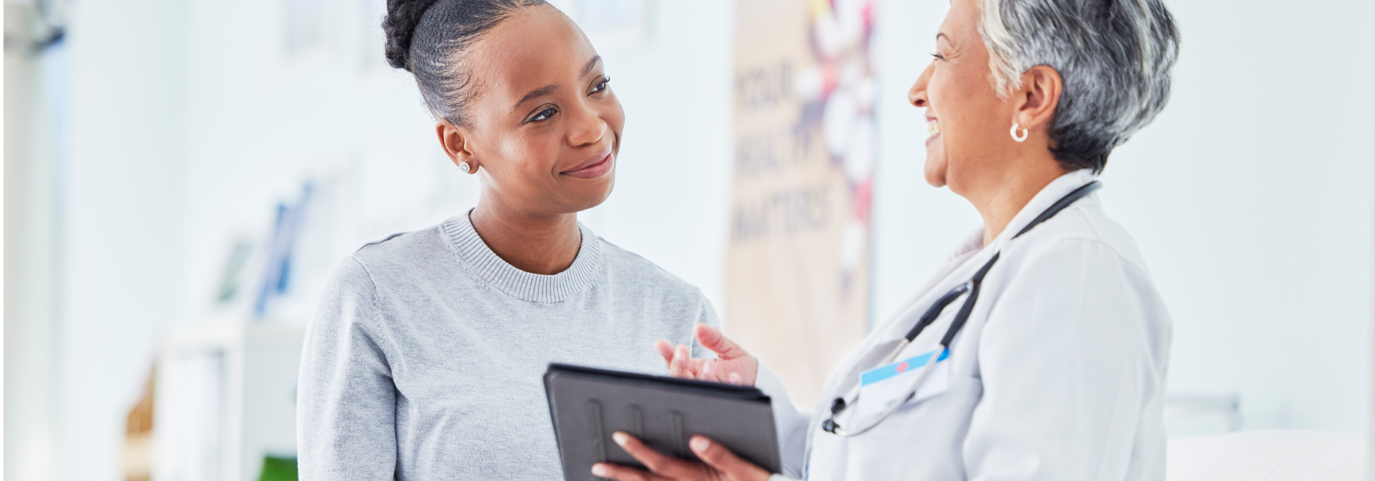 A smiling black woman talking to a smiling older Asian doctor wearing a white lab coat and stethoscope holding a tablet.