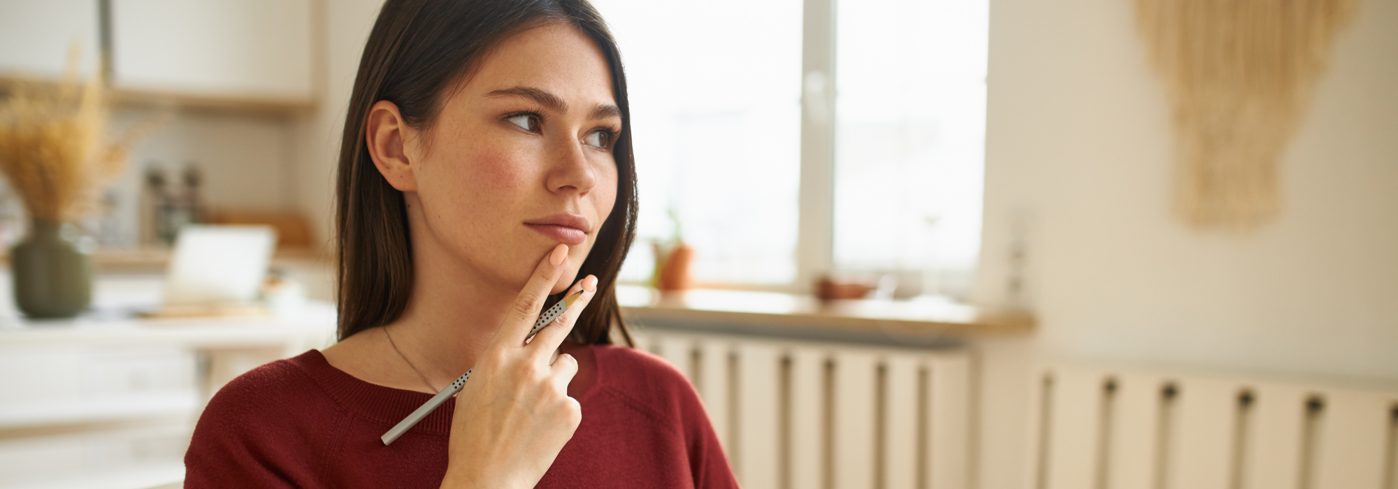 A young woman looking thoughtful, sat down holding a pen in her hand.
