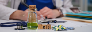 The word 'IBS' on wooden dice on a desk next to a pill blister packet, green liquid in a clear vial, and a stethoscope. A female clinician's hands are in the background, writing something down. She is wearing a white lab coat.