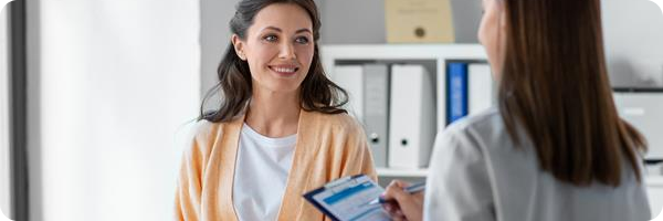 A smiling female patient facing a female doctor who is holding a clipboard and pen. 