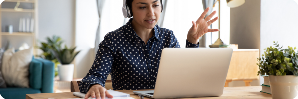 A young professional woman wearing headphones talking to a laptop screen as if on a video call, working from home. 