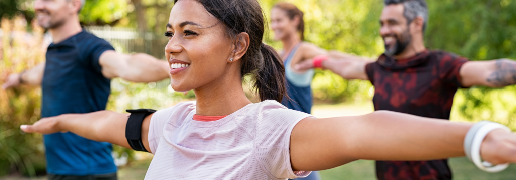 A young smiling woman doing arm stetches outdoors with a group of people.