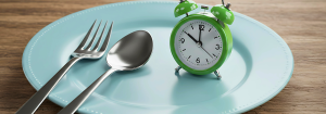 A silver fork and spoon resting on a large dinner plate with a green alarm clock also in the plate showing the time of 10 o'clock.