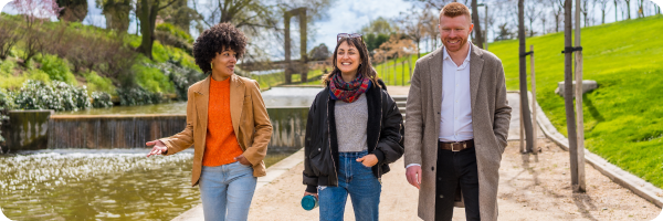 A young black woman, young Caucasian woman and young Caucasian man walking by a river with trees and grass in the background. 