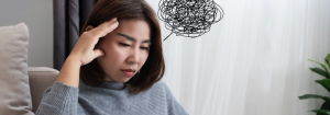 An Asian woman sitting down indoors resting her head in her hand looking pensive with a black scribble thought bubble coming from her head to represent stress and the impact of the brain on the gut and IBS.