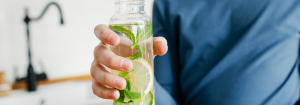 A young Caucasian man's hand holding a glass of water containing lemon slices and green leaves to represent the topic of hydration and IBS.