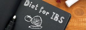 A notebook with black paper open on a wooden desk with the words 'Diet for IBS' written in white above drawings of vegetables to represent different dietary strategies for managing IBS and symptom relief.