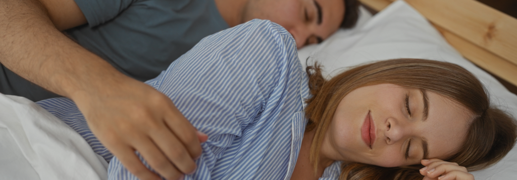 A sleeping young Caucasian man with his arm over a young Caucasian woman sleeping on her side, both in bed to represent the topic of sleep and IBS.