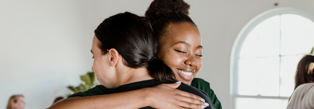 An image of a young smiling black woman hugging a Caucasian woman to represent supporting someone with IBS.